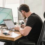 Entrepreneur sitting at work desk using a laptop and a monitor; entrepreneurs long desk hours