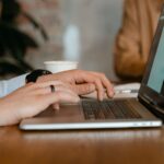 A person typing on a laptop at a wooden table in a modern office setting; ai side hustles