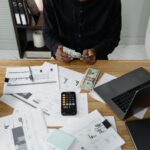 A person counts dollar bills at a desk with a laptop, papers, and calculator; young entrepreneurs personal finance