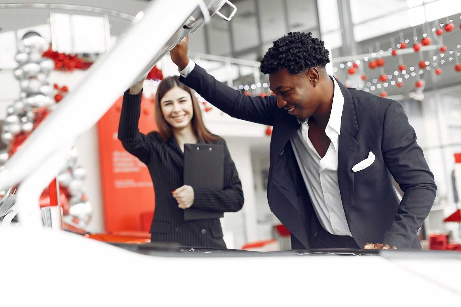 Positive smiling diverse stylish man and female dealer checking under car motor hood and smiling while choosing new car in car showroom; agentic commerce